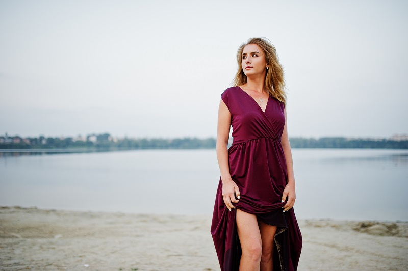 Convertible Dresses Blonde sensual barefoot woman in red marsala dress posing against lake on sand.
