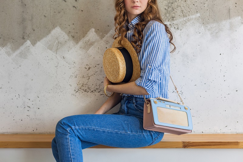Flap Bags: Chic and Subtle young attractive stylish woman sitting at wall background, holding straw hat, handbag, wearing blue cotton shirt, jeans, casual outfit, hipster vintage style, spring summer fashion trend, accessories